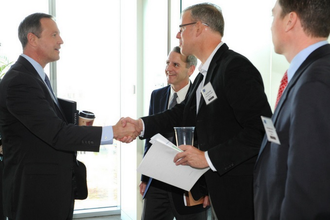 Four men in business attire, two of them shaking hands and smiling, while the other two converse in the background, in a well-lit room with large windows.