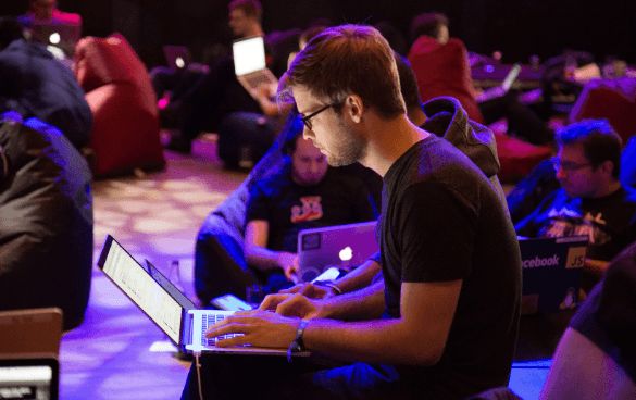 A man with glasses working on a laptop in the foreground with other individuals in the background also using laptops, possibly at a tech event or hackathon.