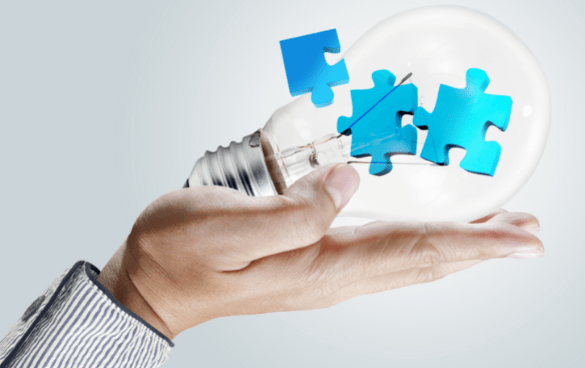 A hand holding a clear light bulb with two blue puzzle pieces inside it, symbolizing creativity and problem-solving.