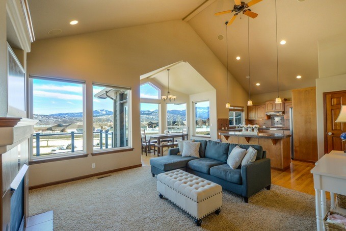 Spacious living room with high vaulted ceilings, large windows with a hilly view, a blue sectional sofa, white ottoman, open-plan kitchen, and a dining area.