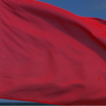 A plain red flag fluttering against a clear blue sky.