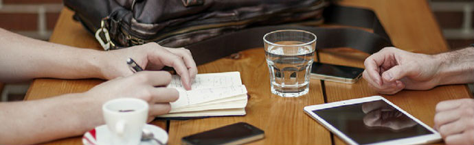 Two people at a wooden table with one writing in a notebook and the other holding a stylus by a tablet, accompanied by a coffee cup, a glass of water, smartphones, and a bag.