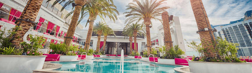 Outdoor swimming pool at a modern resort with pink accents, surrounded by palm trees and lounge chairs, with reflective glass buildings and a partly cloudy sky.