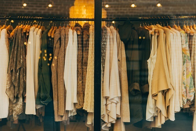 A selection of clothing in earthy tones displayed on a rack in front of a store window with a brick wall and hanging lights in the background.