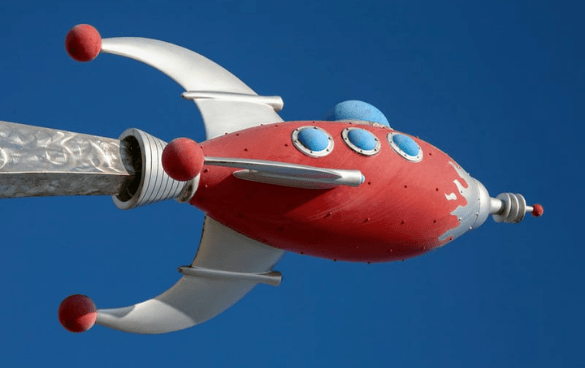 Stylized red and white spaceship model with blue circular windows and red spherical accents against a clear blue sky.