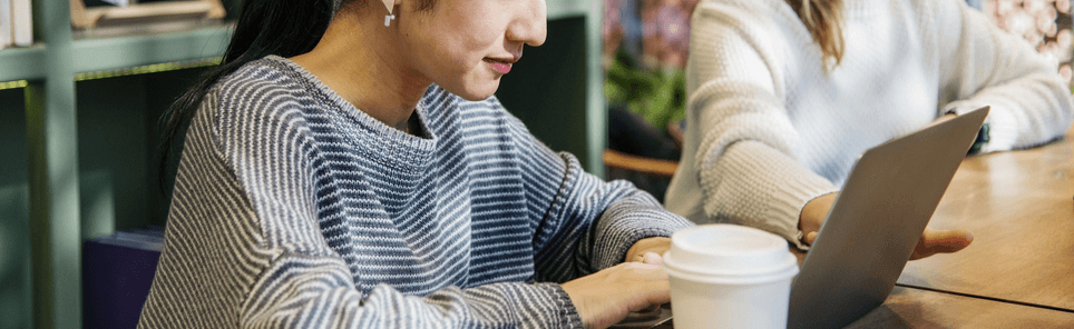 Person in a striped sweater using a tablet with a stylus at a table with a coffee cup in front, another person in a white sweater in the background.