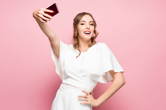 A smiling young woman in a white dress taking a selfie with her smartphone against a pink background.