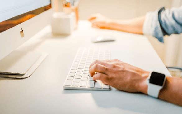 Person typing on a white keyboard at a desk with an iMac, a white mouse, and a cup, wearing a smartwatch and a striped shirt with rolled-up sleeves.