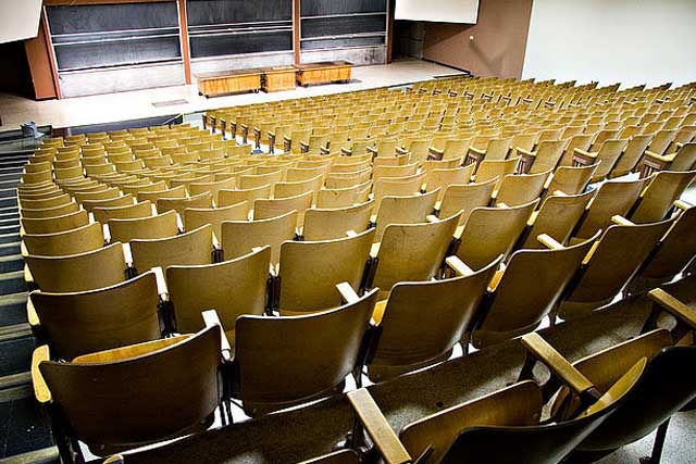 Empty lecture hall with rows of brown chairs facing a stage with a blackboard and two wooden podiums.