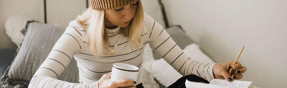 A woman in a beanie and striped shirt holding a mug and writing in a notebook.