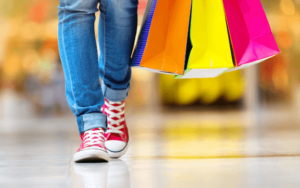 Person wearing blue jeans and red sneakers carrying colorful shopping bags, walking in a blurred commercial setting.