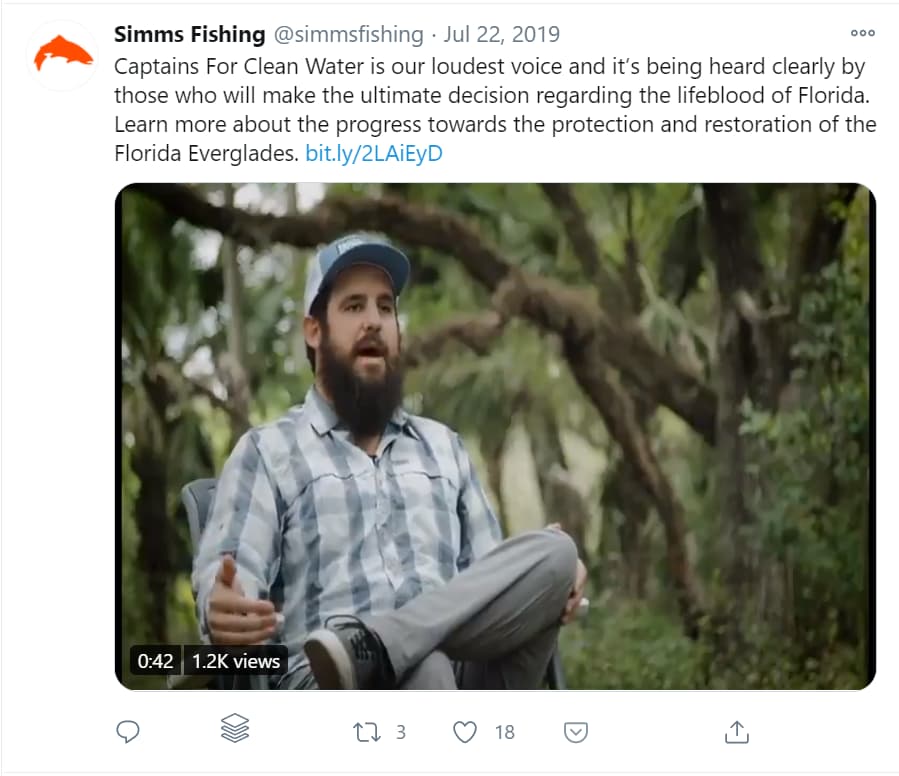 A man with a beard wearing a blue cap and a plaid shirt is sitting outdoors with trees and foliage in the background, appearing to be speaking or gesturing.