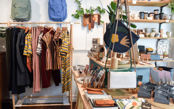 Interior of a boutique shop with a clothing rack on the left and shelves with bags, notebooks, wallets, and ceramic pottery on the right, complemented by hanging plants.