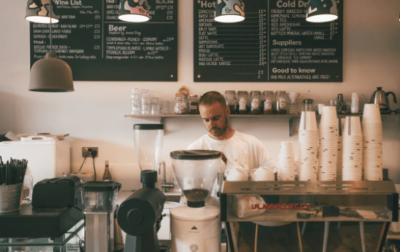 A barista attentively working at a coffee bar with a grinder and disposable cups, with a menu board in the background listing beverages and prices in a cozy coffee shop.