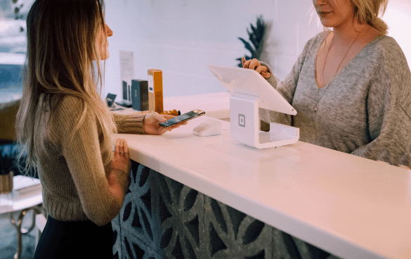 Two women at a checkout counter with one holding a smartphone, possibly making a payment, and the other behind the counter interacting with her, with a point-of-sale system in between them.