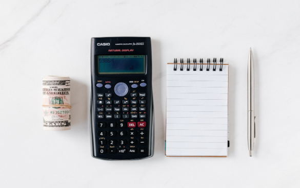 A roll of US dollar coins, a Casio scientific calculator, a small lined notepad, and a pen on a white marble surface.