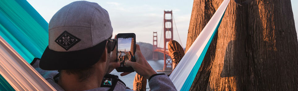 Person in a hammock capturing the Golden Gate Bridge on a smartphone between two tree trunks.