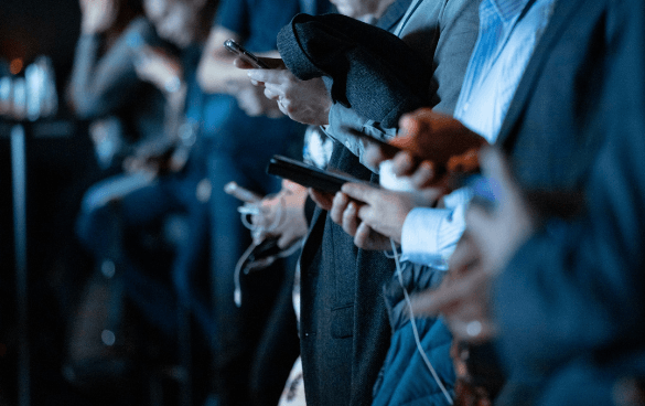 A row of people seated closely, each engrossed in their own smartphone, in a dimly lit event space.