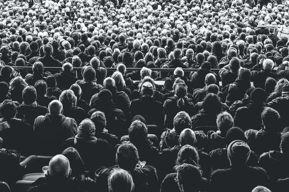 Monochrome image of a densely packed audience viewed from behind in a stadium or auditorium setting.