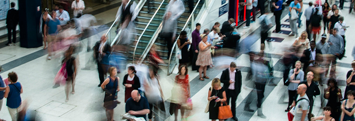 A long exposure shot of a busy indoor transit area with blurred figures in motion and some individuals standing still, conveying the hustle of a commuting environment.