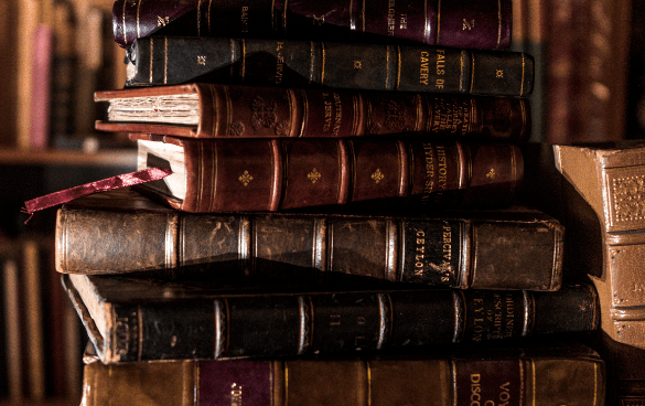 A stack of antique hardcover books with gold lettering on the spines, placed against a blurred background suggestive of a library.