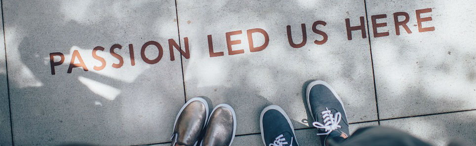 Two pairs of feet wearing different shoes standing on a concrete surface with the inscription 'PASSION LED US HERE' and shadows cast across the ground.