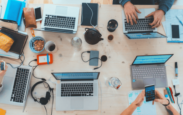 Overhead view of a busy workspace with multiple people using laptops, smartphones, and taking notes, surrounded by personal items like headphones and a coffee cup.