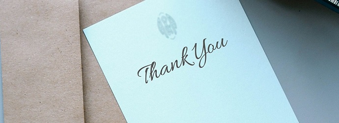 A 'Thank You' card with cursive handwriting on a brown paper surface with a pen partially visible on the right.