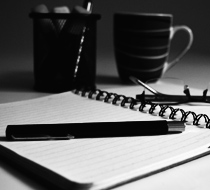 Black and white photo of a desk with an open notebook, a pen, a cup with writing utensils, and a coffee mug.