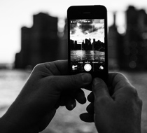 A person's hands holding a smartphone, taking a photo of a city skyline at dusk through the phone's camera interface, with the cityscape visible on the phone's screen.