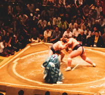 Two sumo wrestlers facing off in a dohyo with a referee observing, surrounded by an attentive audience.