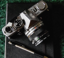 A vintage silver film camera with dials and a lens, alongside a black pen with a silver clip, on a dark surface.