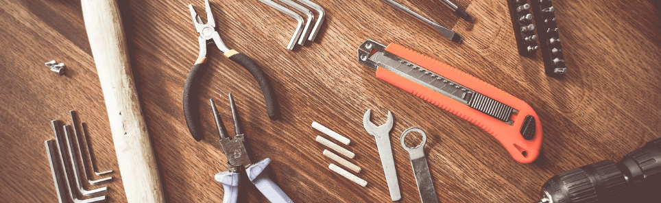 A variety of tools including pliers, wrenches, a box cutter, hex keys, and a drill, along with screws and bits, spread out on a wooden surface.