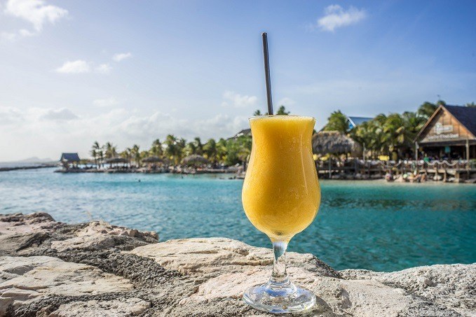 A frozen yellow beverage in a glass with a straw on a rocky ledge, overlooking a tropical beach resort with thatched-roof buildings and clear blue waters under a sunny sky.