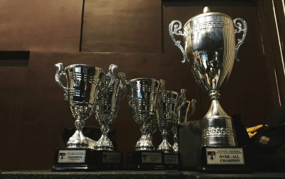 A collection of five shiny trophies on a shelf, with the largest on the right labeled 'OVER-ALL CHAMPION' and four smaller ones to the left.