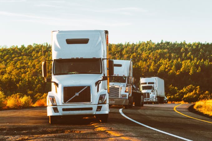 A convoy of white semi-trucks on a curving road at sunrise or sunset with a backdrop of green trees and an orange sky.