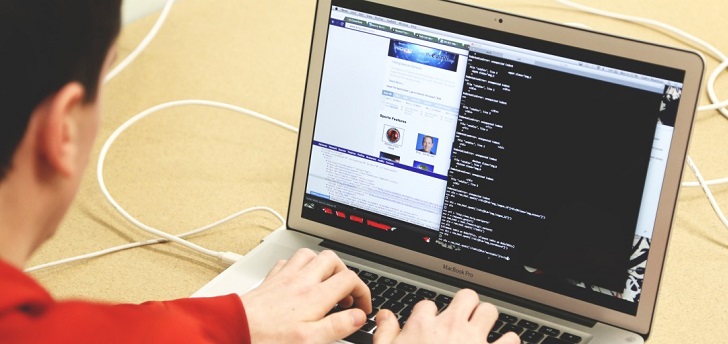 Person in a red shirt using a MacBook Pro on a desk with visible code and a web page on the screen.