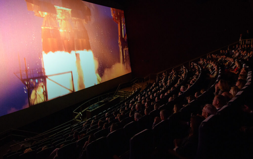 Audience watching a space rocket launch on a large screen in a movie theater.