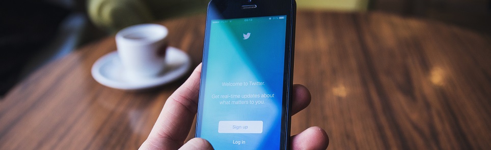 Person holding a smartphone displaying the Twitter login screen with a blurred cup of coffee in the background.