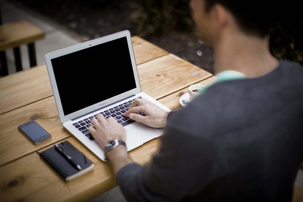 Person from behind working on a laptop with a black screen at a wooden table, with a smartphone, closed notebook with pen, and a cup of coffee beside them.