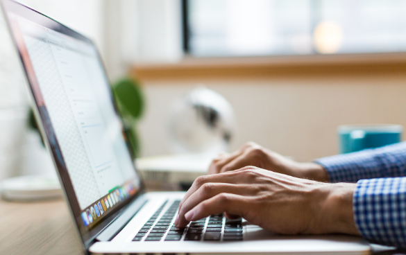 Close-up of a person's hands typing on a laptop keyboard with a blurred background featuring a cup and a decorative item.