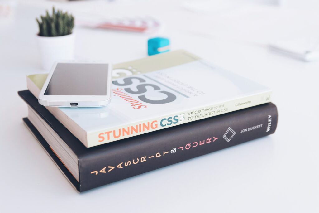 A white smartphone on top of two web development books titled 'Stunning CSS3' and 'HTML & CSS' on a white desk, with a potted cactus, a blue eraser, and a pen in the background.