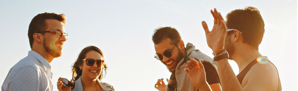 Four young adults laughing and enjoying a conversation in the sunlight, three of them wearing sunglasses.