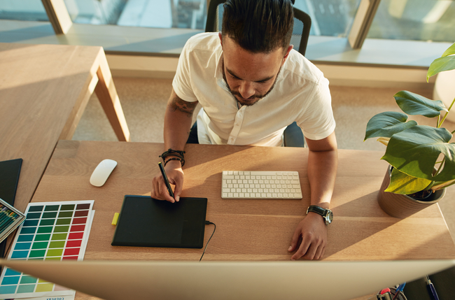 A man with a man bun hairstyle working on a graphic tablet at a wooden desk, with a keyboard, mouse, color swatches, and a potted plant, in a bright modern office.