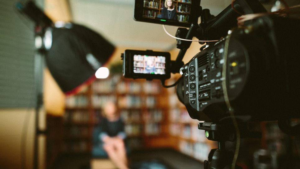 A professional video camera setup recording an out-of-focus person sitting in front of a bookshelf, with a softbox light on the left and the camera's viewfinder showing the subject.