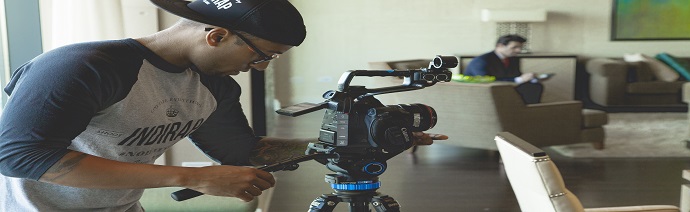 A person adjusting a professional video camera on a tripod with another person sitting in the background.