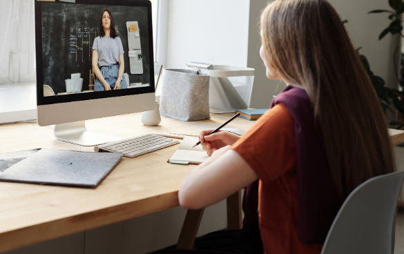 Person attending an online class or meeting from a home office, taking notes while watching a presenter on the computer screen.