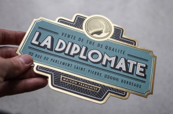 A person holding a decorative label for 'La Diplomate' with text indicating quality sales and a French address in Bordeaux, designed in dark blue, light blue, and gold with a wheat sheaf graphic.