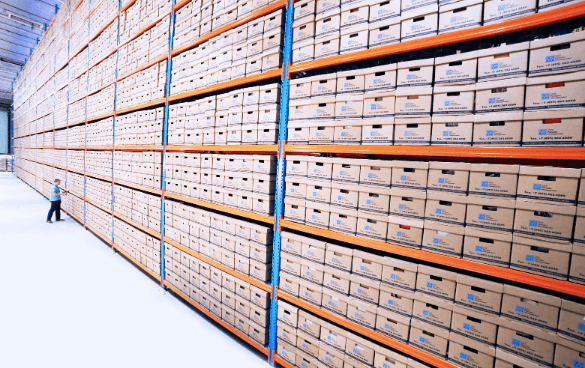 A person in a high-visibility vest walks beside towering shelves filled with labeled boxes in a warehouse.