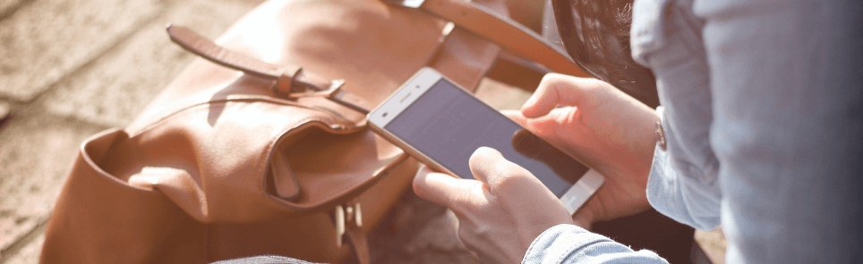 Close-up of a person's hands using a smartphone beside a brown leather bag, with sunlight casting shadows.